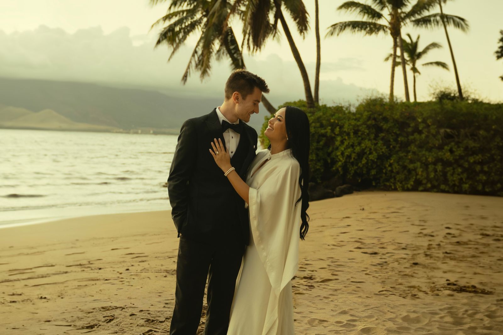 Alex and Megan Colhoun on a beach with palm trees at sunset, Alex in a tuxedo and Megan in a white dress