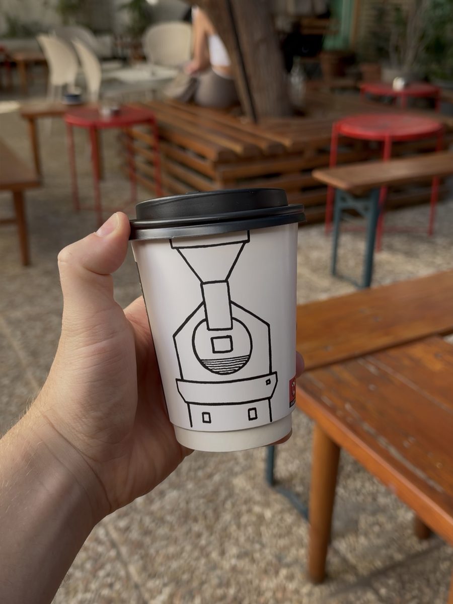 A Paul's Coffee Roasters takeaway cup with a coffee roaster drawing, held in the courtyard