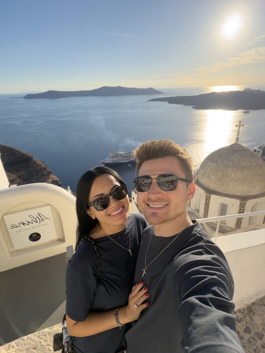 Alex and Megan Colhoun taking a selfie at the Athina Aperto Suites viewpoint in Fira, with the caldera and a church dome behind