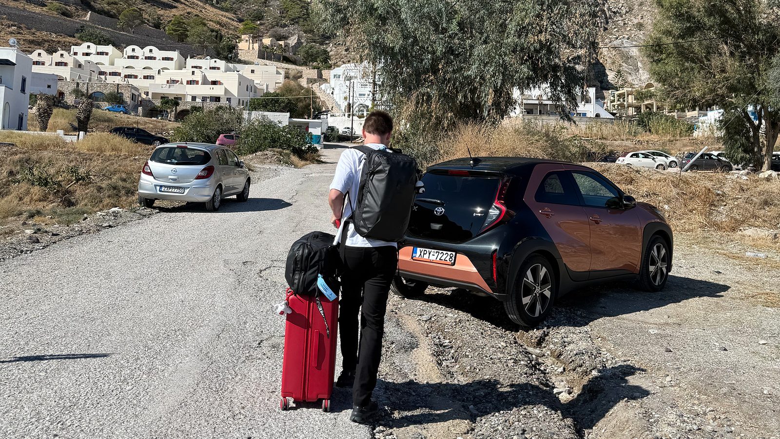 Alex walking with luggage to a Sixt rental car on a road in Santorini