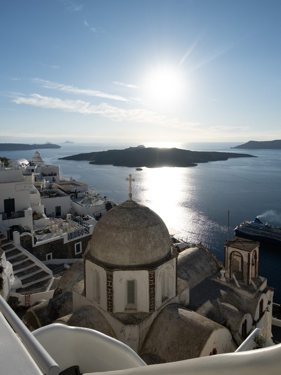Church dome with a cross above the Santorini caldera, with the volcano, the sun over the sea and a cruise ship, from a viewpoint in Fira