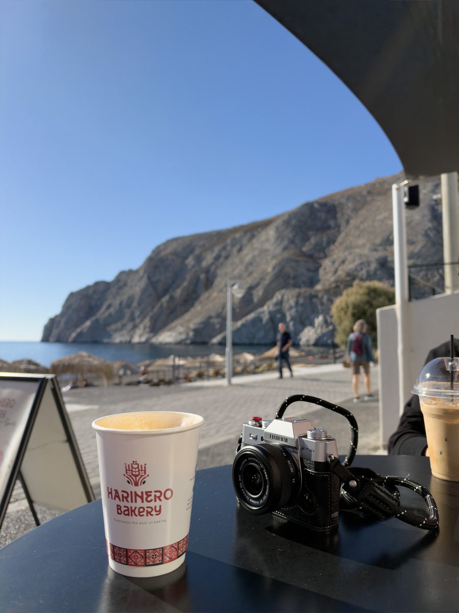 Harinero Bakery latte and a Fujifilm camera on a table facing Kamari beach and the Mesa Vouno cliff