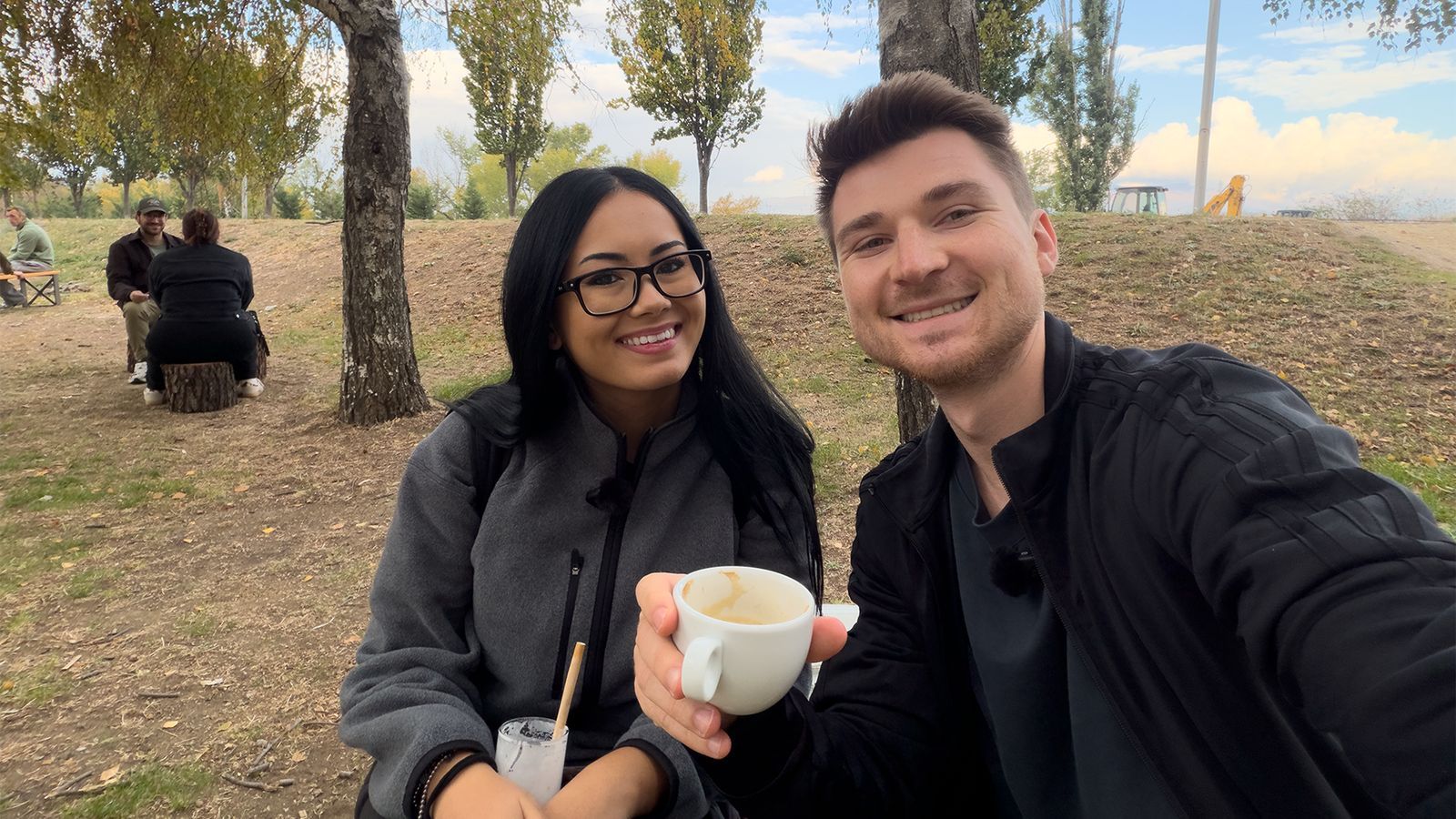 Alex and Megan having coffee at an outdoor table under trees in Skopje